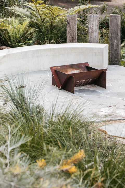 Rust-colored corten steel fire pit with crazy paving in a garden setting with greenery and concrete.